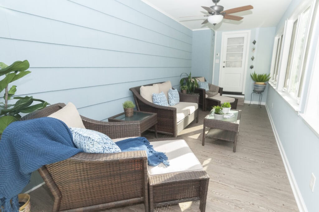 A bright sunroom with blue walls, wicker chairs with cushions, blue throw pillows and blankets, glass-top tables, potted plants, a ceiling fan, and large windows letting in natural light.