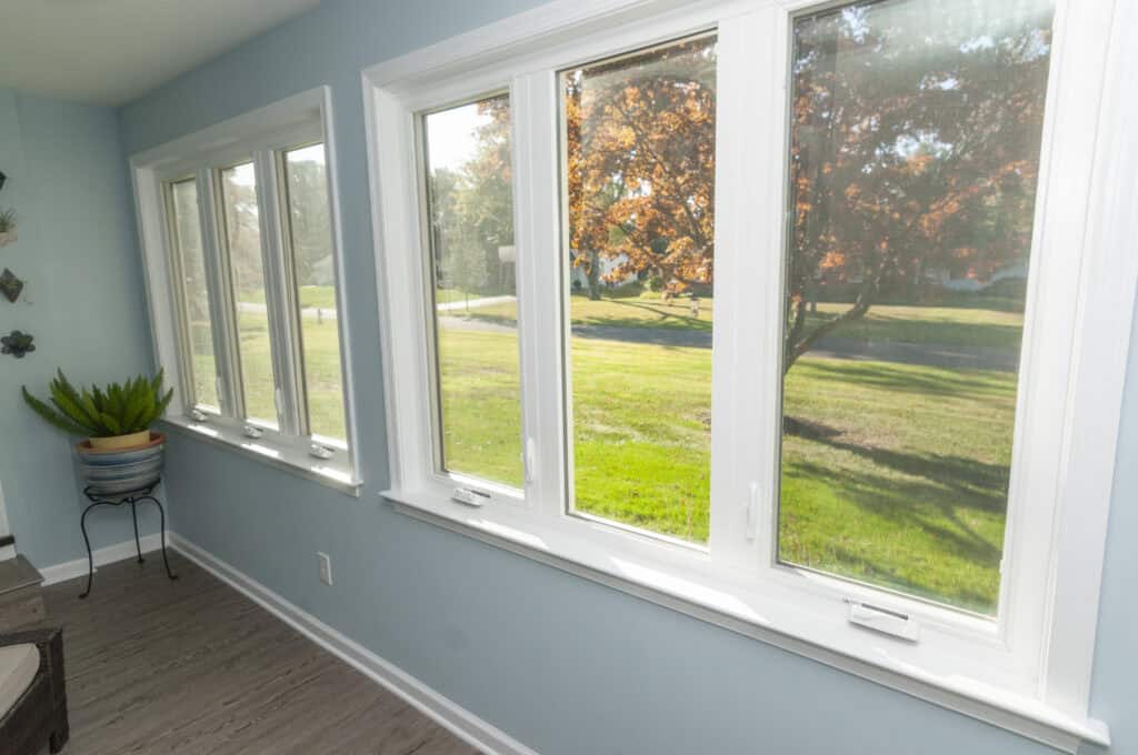 Large white-framed windows in a light blue room overlook a green lawn and trees with orange leaves, letting in bright sunlight. A potted plant sits on a small stand near the corner.
