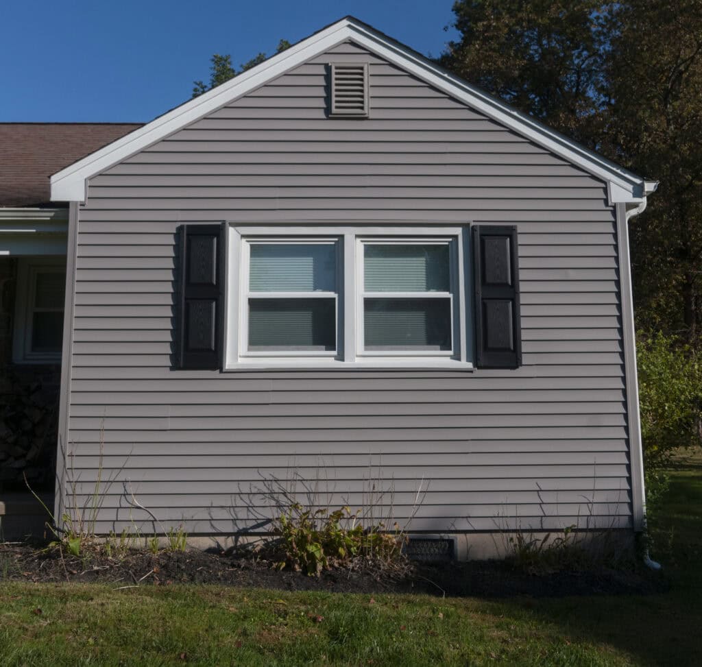 The image shows the side of a gray house with white trim, one double window with closed blinds, black shutters, and a small garden bed with sparse plants beneath the window.