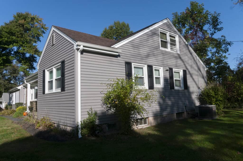 A gray, single-family house with white trim sits on a grassy lawn. There are small shrubs and plants along the side, black shutters on the windows, and trees in the background under a clear blue sky.