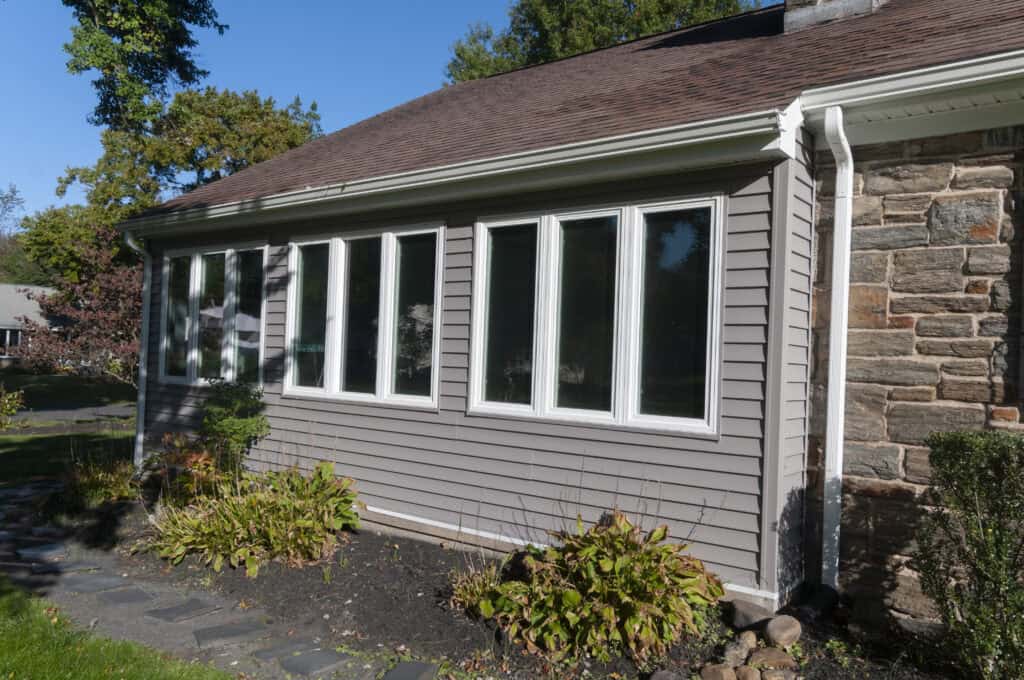 A house with gray siding and six tall rectangular windows, next to a stone wall. There are plants and a stone path in the yard under a clear blue sky.