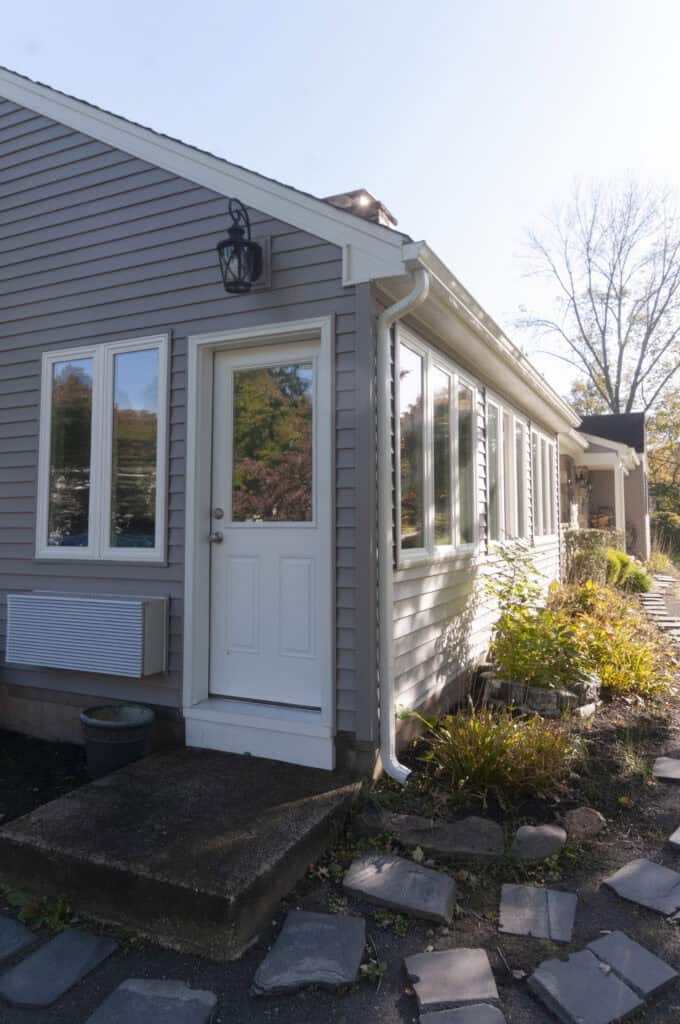 A beige house with white trim, a white door with glass, rectangular windows, and a stone path leading to the entrance. Plants and shrubs line the walkway, and sunlight shines on the scene.