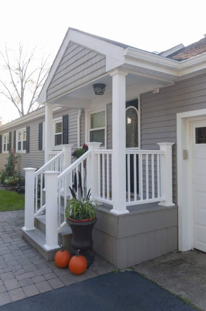 A small porch with white railings and steps leads to a gray house. Three orange pumpkins and a tall planter with green plants decorate the entrance, giving a seasonal autumn feel.