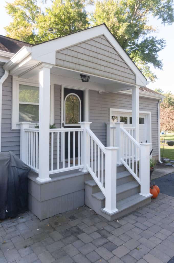 A small, gray house with a white front porch, white railings, and steps leading up to a glass front door. There are trees in the background and a pumpkin on the right side of the steps.