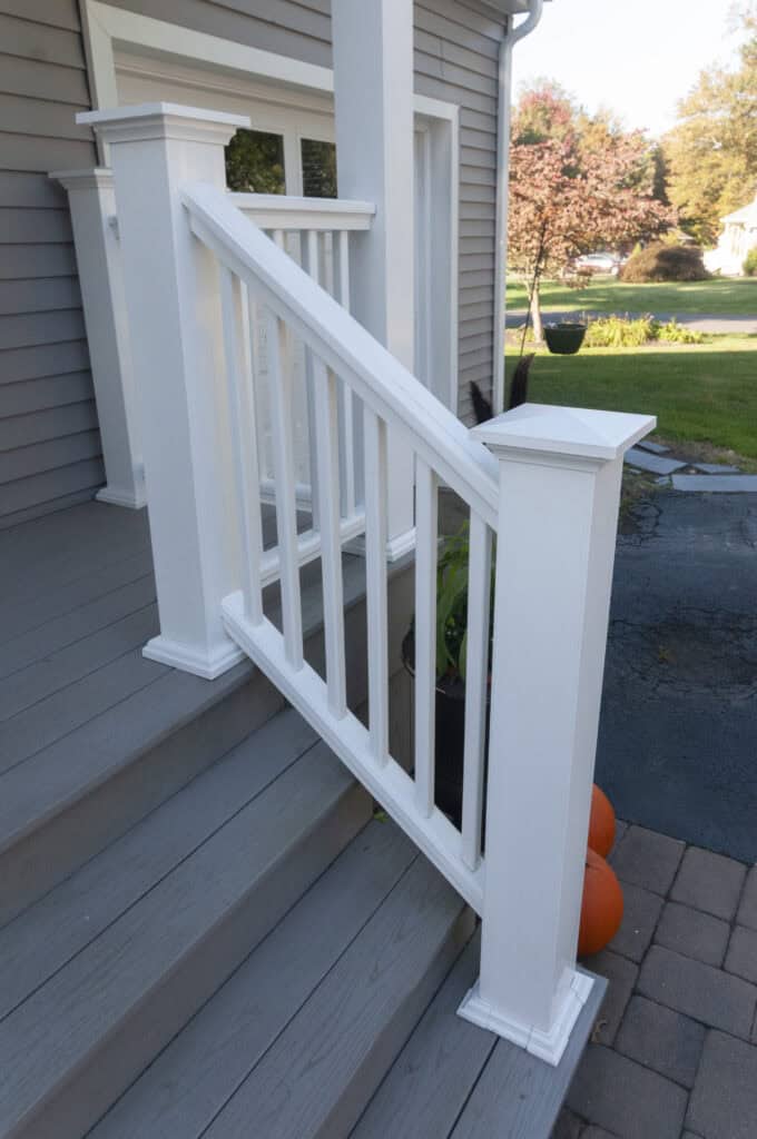 White wooden railing on gray porch steps of a house, with pumpkins at the base and a driveway, green lawn, and trees visible in the background.