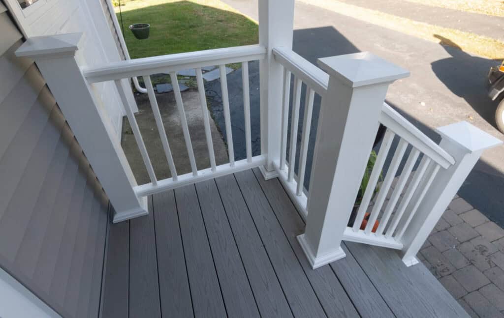A small porch with gray wooden flooring and white railings, leading down three steps to a paved walkway. The porch is attached to a light gray house and overlooks a driveway and some grass.