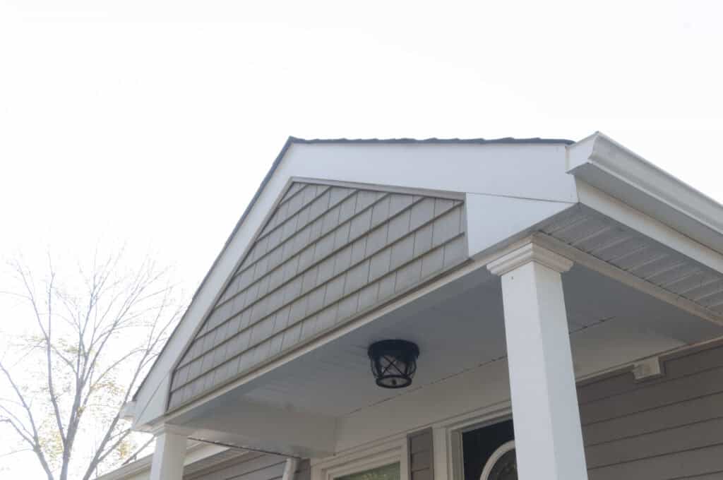 A close-up view of the front porch roof of a house, featuring a triangular gable, shingle siding, white columns, and a black outdoor light fixture underneath. A tree with bare branches is visible in the background.
