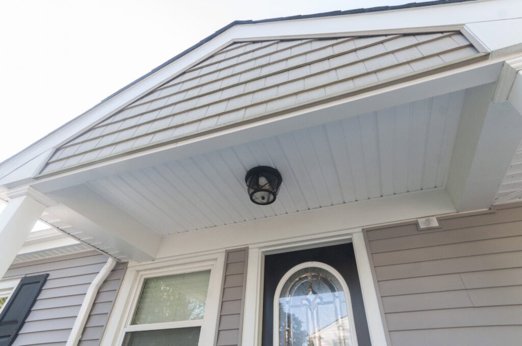 Close-up view of a house entrance with beige siding, a white covered porch ceiling, a black outdoor light fixture, a decorative glass front door, and a window.