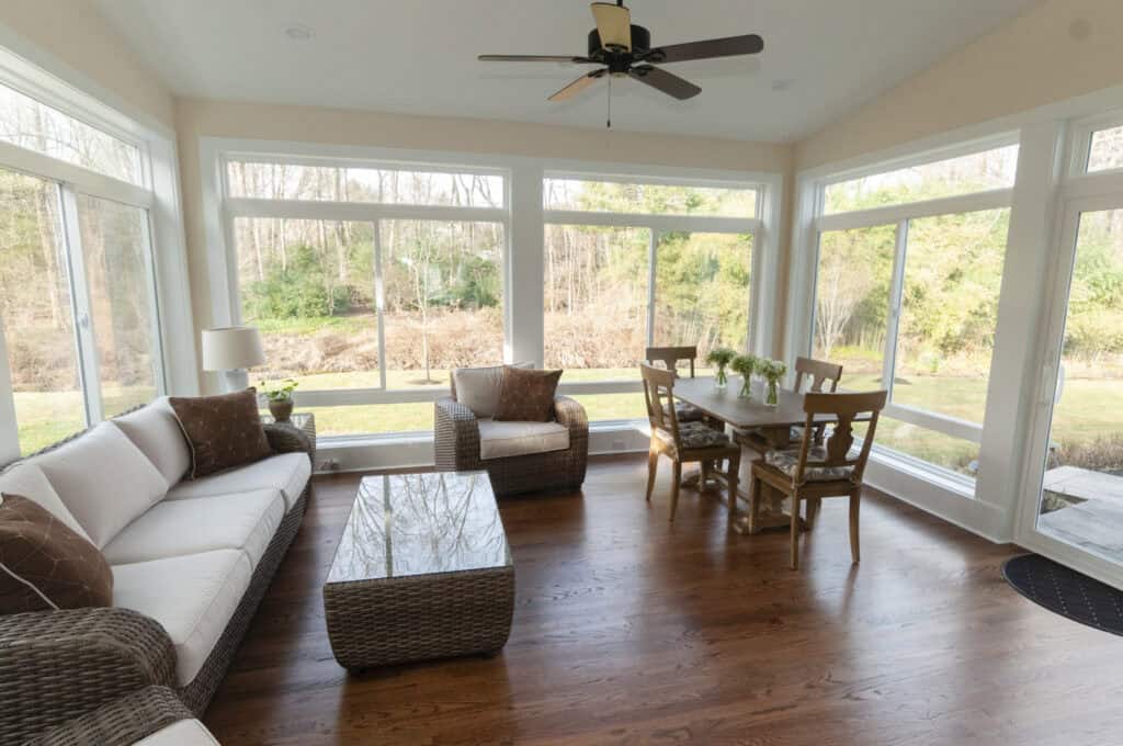 A bright sunroom with large windows, wicker furniture including a sofa, armchair, and coffee table, and a wooden dining table with four chairs. The room overlooks a grassy outdoor area with trees.