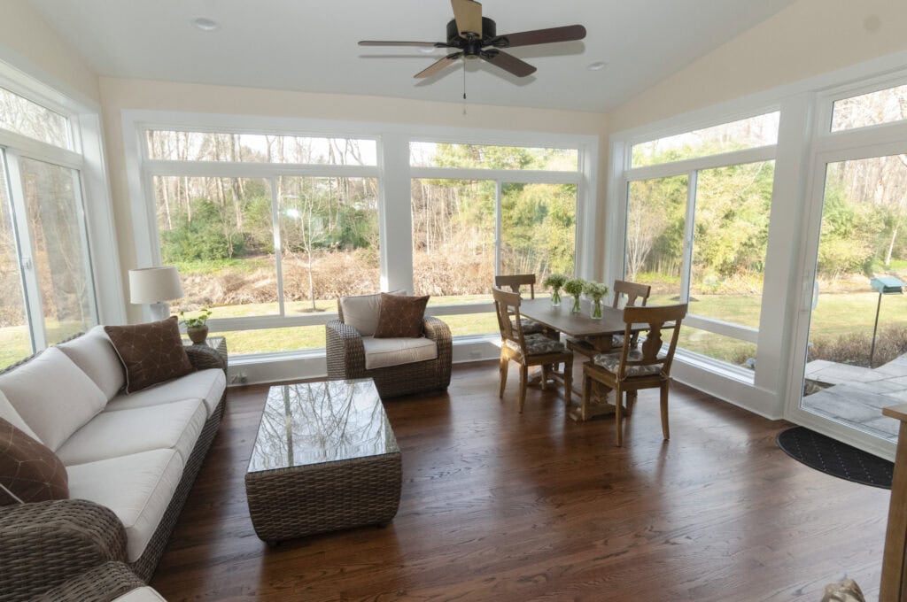 A bright sunroom with large windows, hardwood floors, wicker furniture, a sofa, armchairs, a wooden dining table with chairs, and green plants outside. Natural light fills the cozy, inviting space.