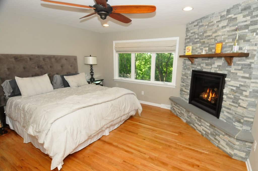 A cozy bedroom with a large bed, light bedding, a lamp, hardwood floors, a ceiling fan, and a stone fireplace with a lit fire. A window with a beige shade lets in natural light and greenery outside.