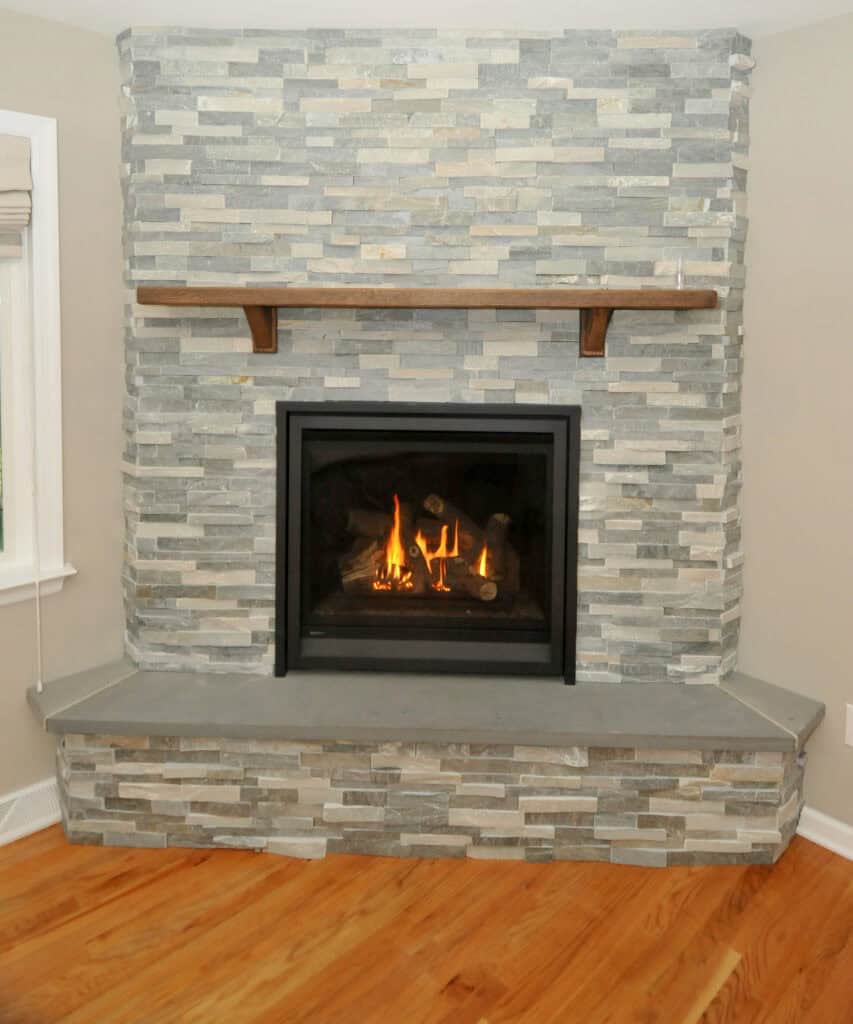 A modern fireplace with a stone surround, wooden mantel, and gas fire burning inside, set against a beige wall with hardwood flooring and a window to the left.