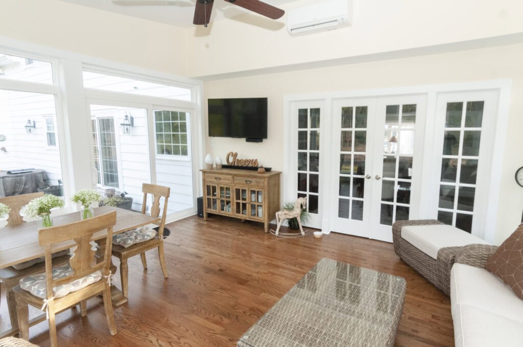 Bright sunroom with wooden floors, dining table with chairs, a TV mounted above a cabinet, glass French doors, and large windows letting in natural light. Cozy seating and decorative plants add warmth to the space.