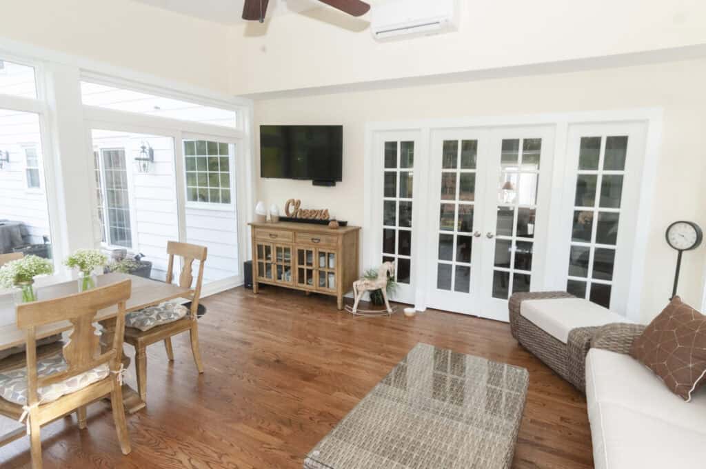 Bright sunroom with hardwood floors, French doors, a wicker sofa, dining table with chairs, glass coffee table, wooden sideboard, wall-mounted TV, houseplants, and large windows letting in natural light.