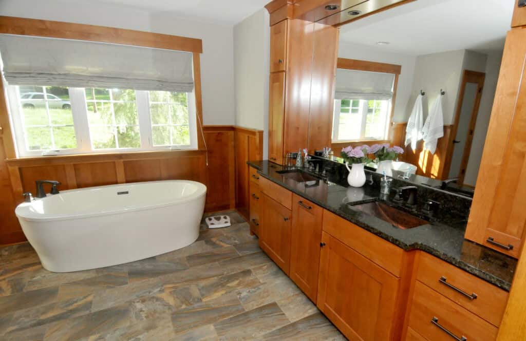 A spacious bathroom with a freestanding bathtub by a window, wooden cabinets with black countertops, a large mirror, and a vase of flowers on the counter. The floor has brownish-gray tiles. Towels hang on the wall.