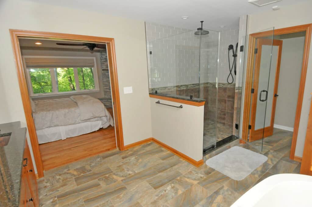 A modern bathroom with tile flooring, a glass-enclosed walk-in shower with a rainfall showerhead, and an entryway leading to a bedroom with a bed and large windows overlooking greenery.