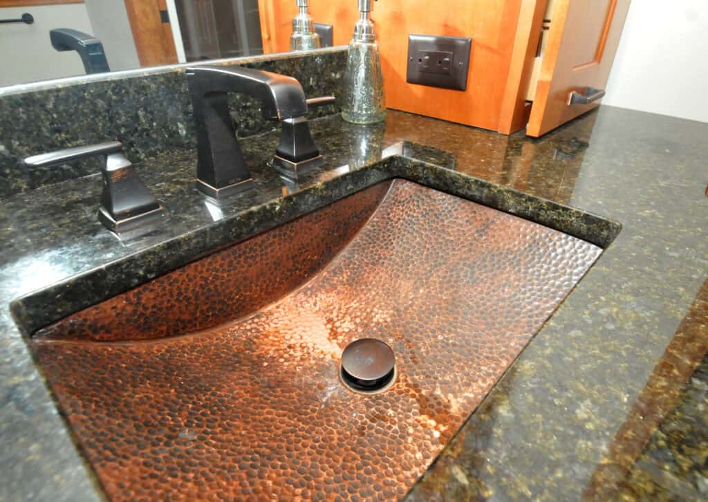 A close-up of a bathroom countertop with a hammered copper sink, black faucet, and soap dispenser. The countertop is dark stone, and a wooden cabinet and mirror are visible in the background.