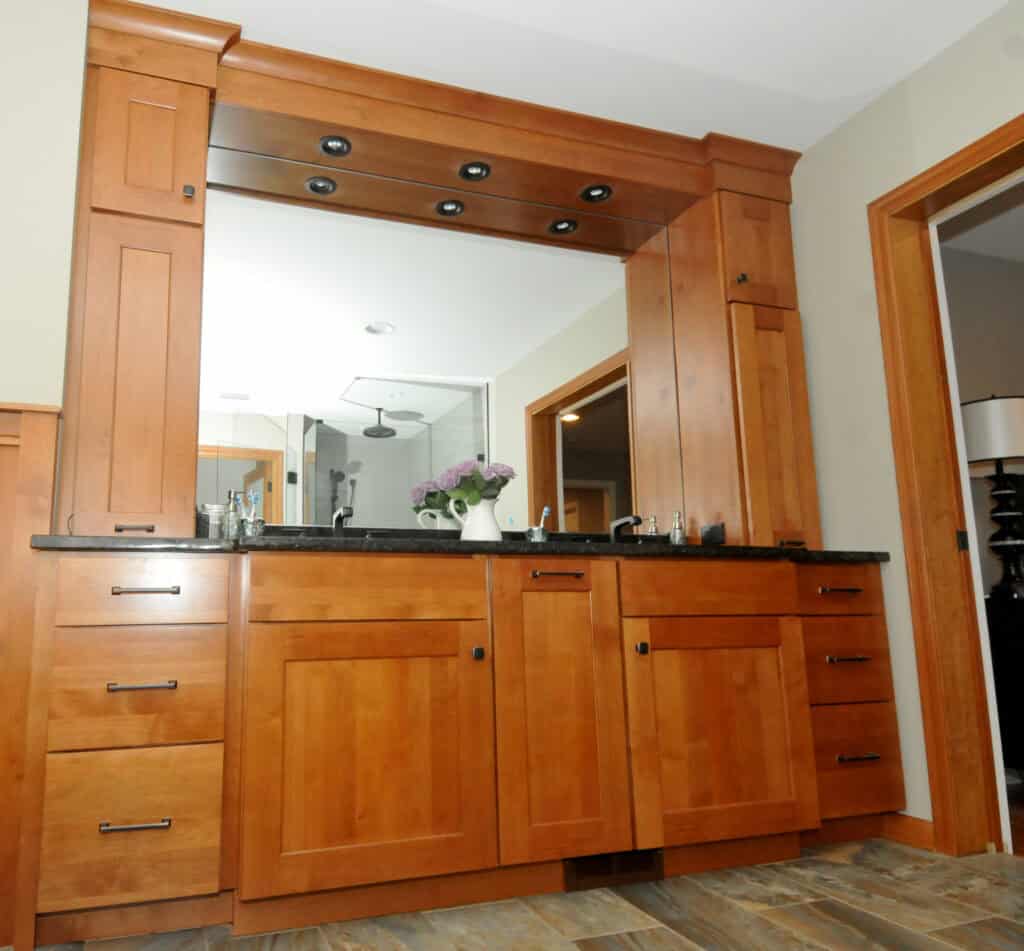 A wooden bathroom vanity with black countertop, double sinks, drawers, and cabinets. Above the sinks is a large mirror framed by wood and spotlights, with a vase of flowers on the counter.