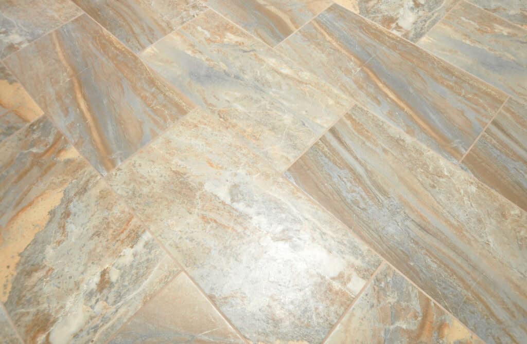 Close-up of a tiled floor with a natural stone pattern in shades of beige, gray, and brown, arranged in an offset grid layout. The surface appears smooth and slightly reflective.