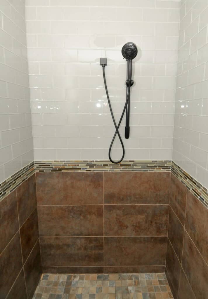 Shower stall with a black handheld showerhead, white subway tile on the upper wall, a horizontal accent strip of small mosaic tiles, brown tiles on the lower wall, and multicolored stone tiles on the floor.
