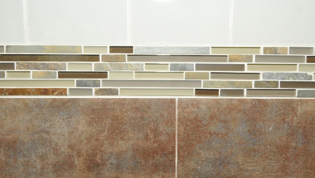 A wall featuring large brown tiles on the bottom, a decorative horizontal strip of small rectangular mosaic tiles in various earth tones in the middle, and plain white tiles on the top.