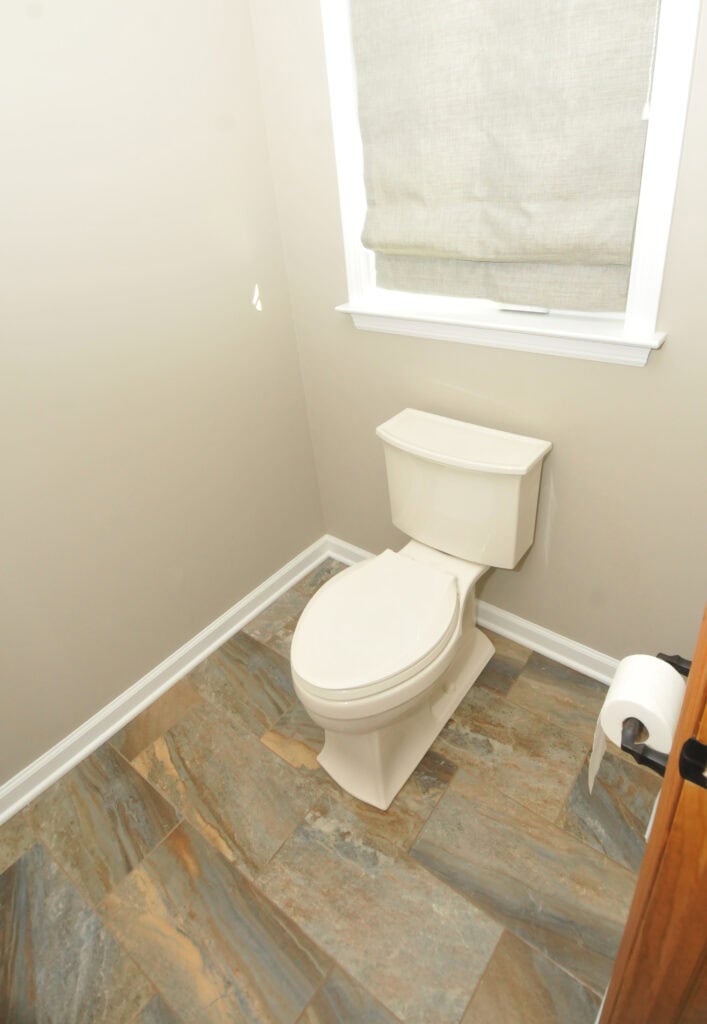 A small bathroom with a white toilet, beige walls, a window with a light fabric shade, and a roll of toilet paper on a holder next to the toilet. The floor has multi-toned brown and gray tiles.