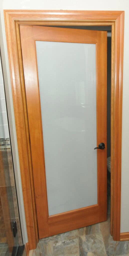 A wooden door with a frosted glass panel in the center, set in a wood frame. The door is slightly open, revealing part of a room with tiled flooring and a glimpse of a bathroom on the left.