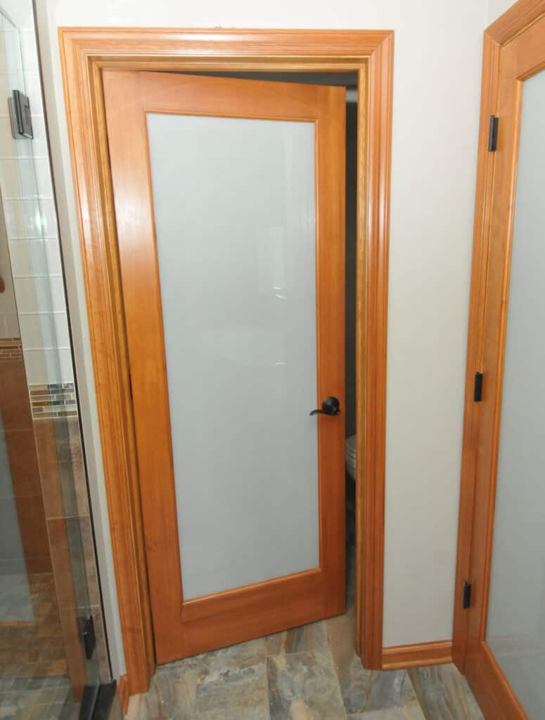 A wooden interior door with a frosted glass panel is partially open, revealing a room inside. The door has a black handle and matches the wooden trim and neutral-colored walls. The floor is tiled with a stone-like pattern.