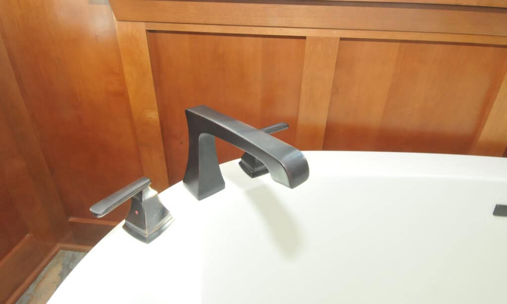 A close-up of a modern black bathtub faucet with two matching handles, set against a white tub and wooden paneled wall.
