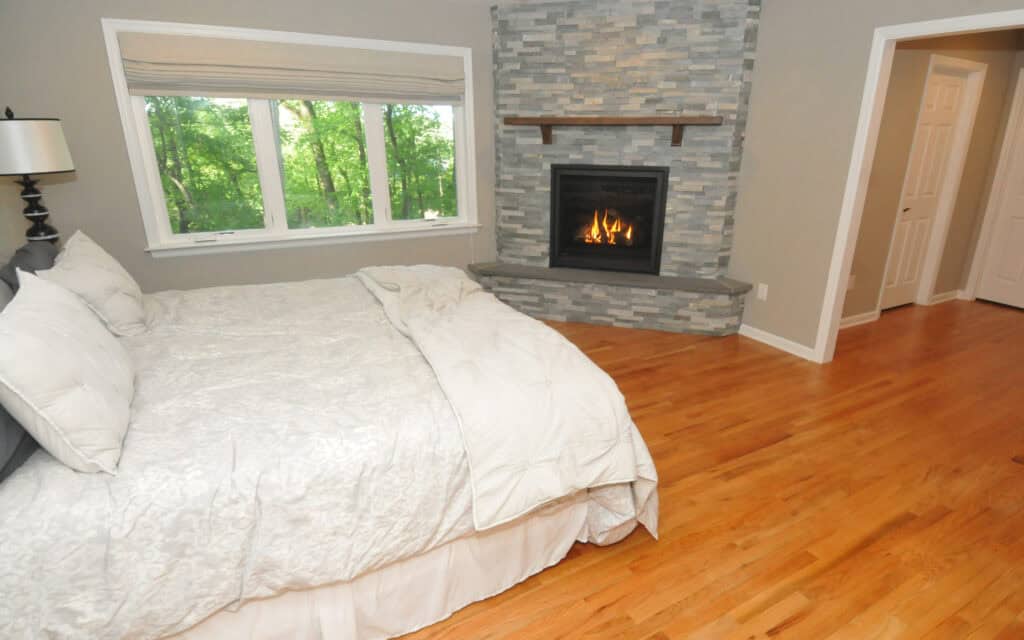 A cozy bedroom with a white bedspread, a window showing green trees, hardwood floors, and a modern fireplace with a gray stone surround below a wooden mantel.