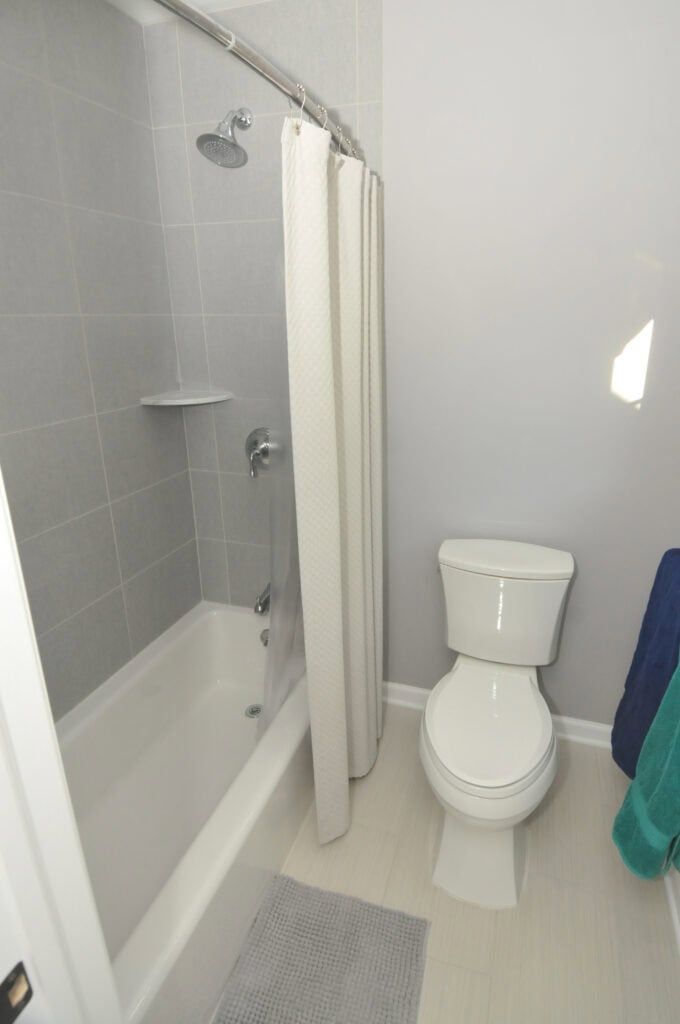 A small bathroom with a white bathtub and shower, gray tiled walls, a white shower curtain, a white toilet, a gray bath mat, and blue and green towels hanging on the wall.