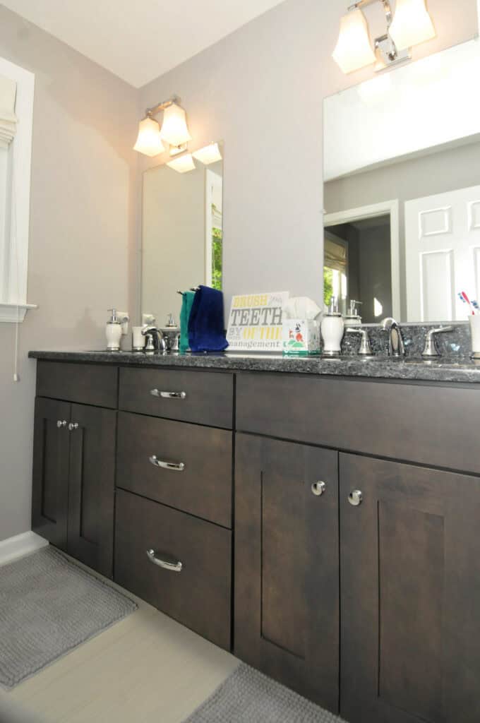 A bathroom with a dark wood double-sink vanity, gray countertop, two mirrors, two light fixtures, and gray rugs. Items like soap dispensers, towels, and a sign about brushing teeth are on the counter.
