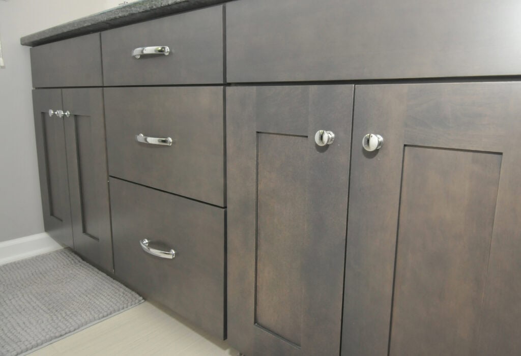 Close-up of modern wooden bathroom cabinets with silver handles and knobs, set against a light-colored tiled floor and a gray textured bath mat.