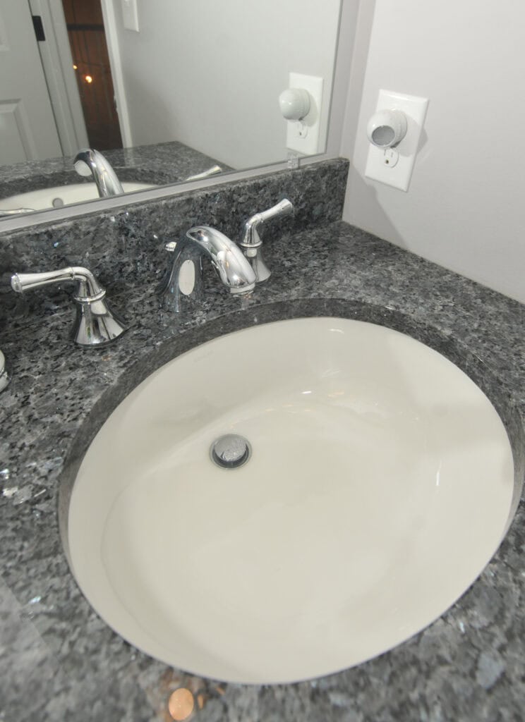 A bathroom sink with a white basin and chrome faucet set in a dark granite countertop, with a mirror and white electrical outlets on the wall behind it.