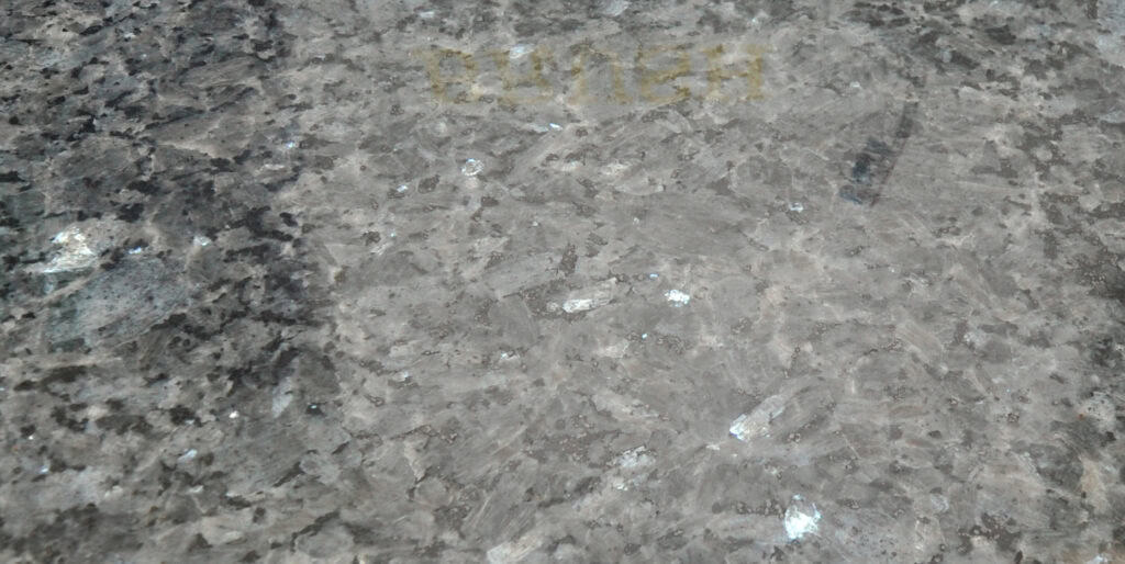 A close-up of a textured, grayish stone surface with scattered white and dark patches. Faint yellow text, partially reading CAUTION, is visible near the top of the image.