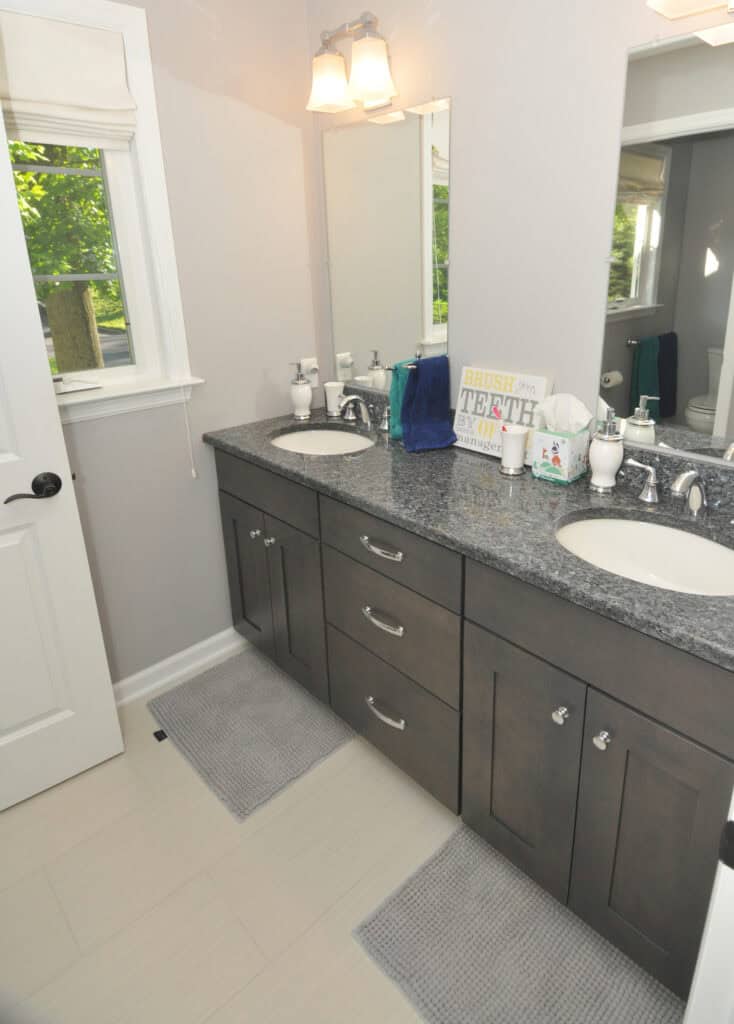 Modern bathroom with double sinks, granite countertop, gray cabinets, two mirrors, a window, and light fixture above. There are two gray rugs on the floor and a sign on the counter that says, Brush your teeth.