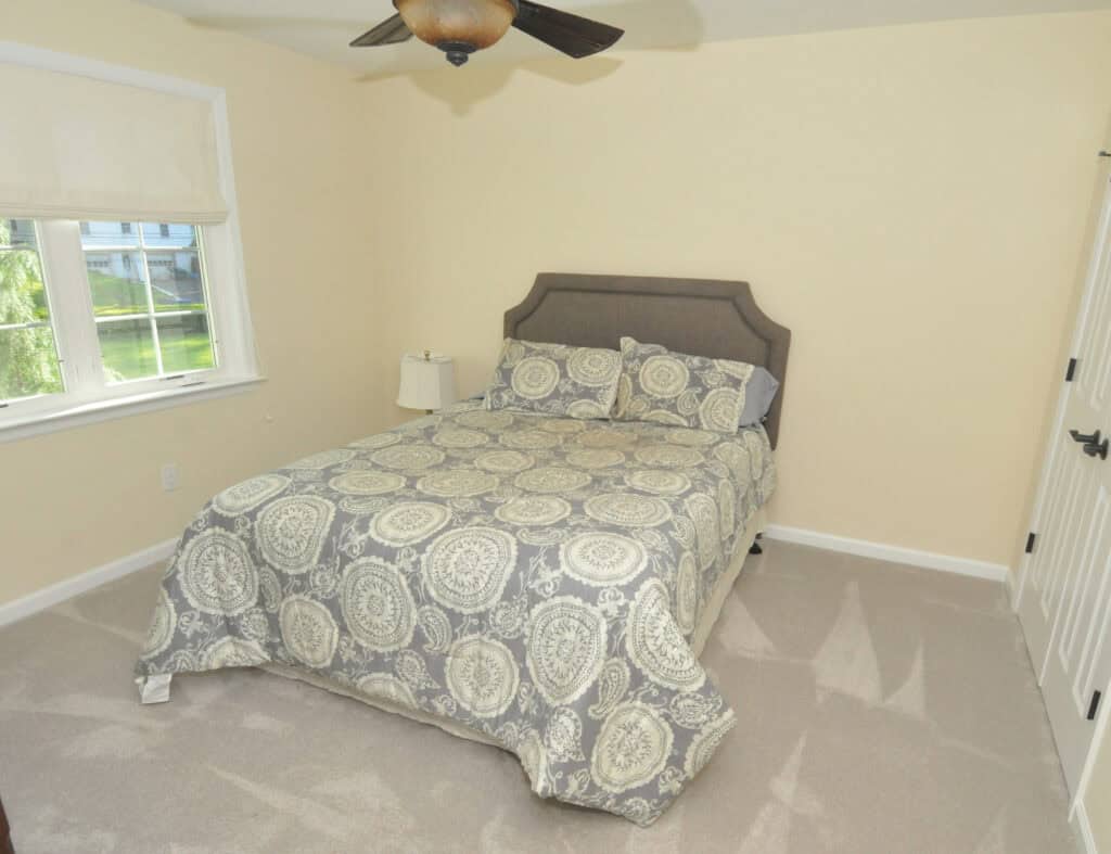 A simple bedroom with a double bed featuring a gray and white patterned comforter, a gray headboard, a bedside table with a lamp, beige walls, a ceiling fan, and a window with a white shade.
