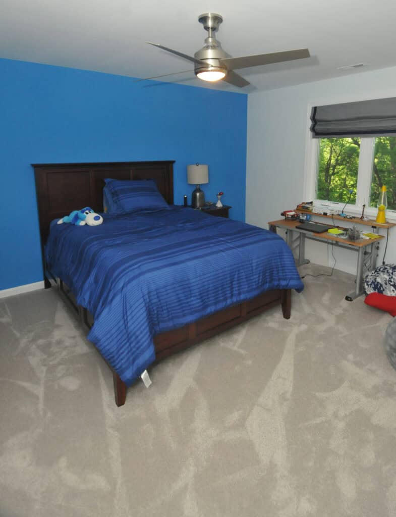 A neatly made bed with blue bedding is centered in a bedroom with a bright blue accent wall. A bedside table, lamp, small stuffed animals, and a desk with various items are visible. A window overlooks greenery outside.