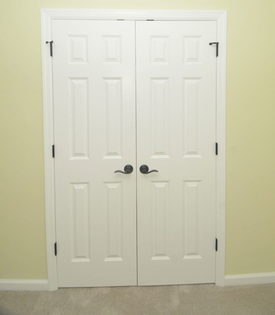 Two white double doors with black handles and hinges, set in a light yellow wall above a beige carpeted floor. The doors have a paneled design and are closed.