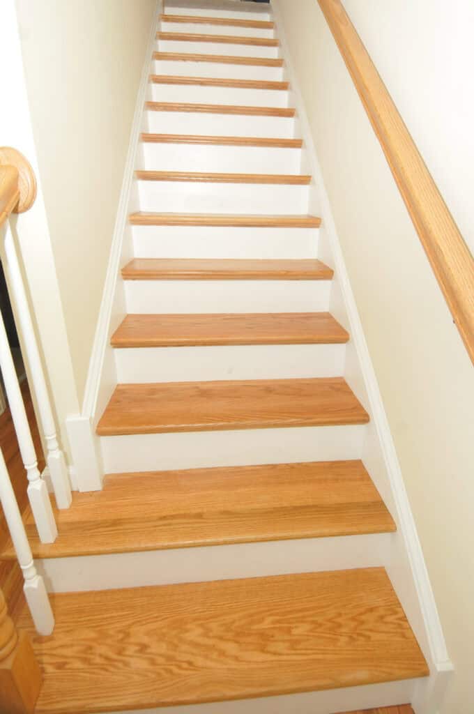 A straight staircase with light wooden steps and white risers, featuring a wooden handrail on the right side and white spindles on the left, set against light-colored walls.
