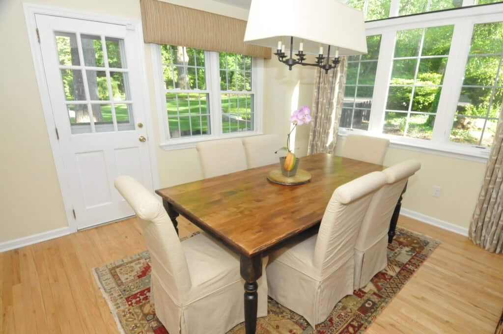 A dining area with a wooden table, four cushioned chairs, a floral centerpiece, a large window with outdoor views, a white door, and a patterned rug on a hardwood floor.