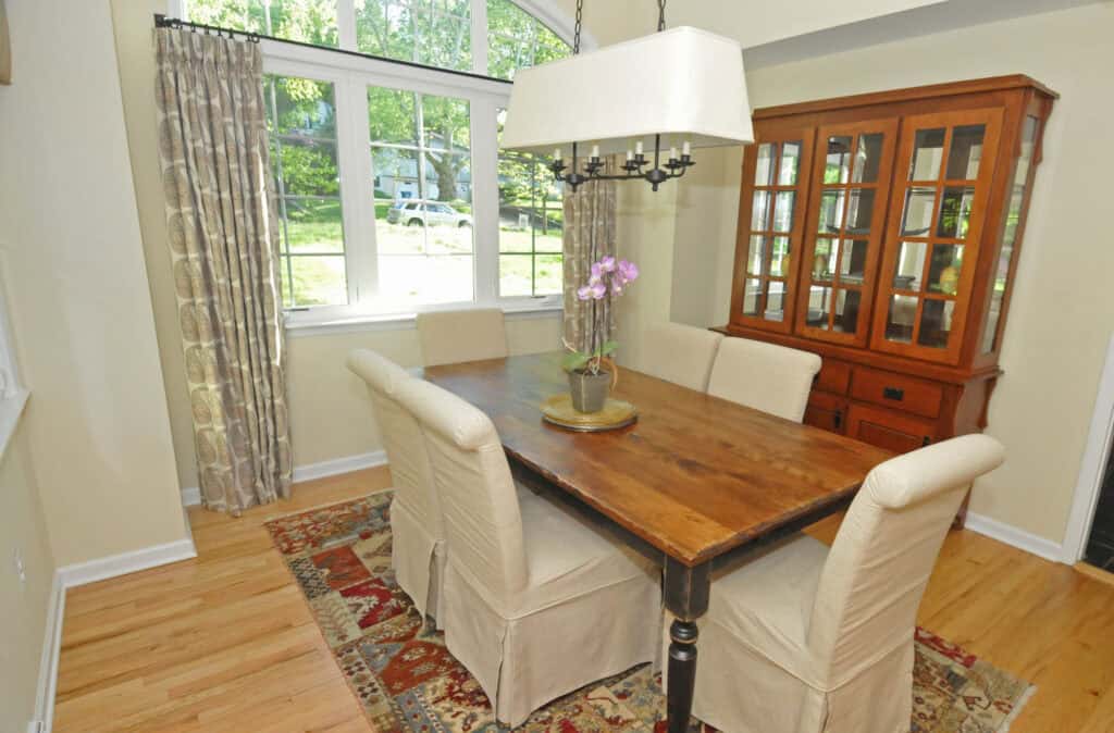 A dining room with a wooden table, four beige upholstered chairs, a patterned rug, a glass-front cabinet, a chandelier, and large windows with curtains letting in natural light. An orchid centerpiece sits on the table.
