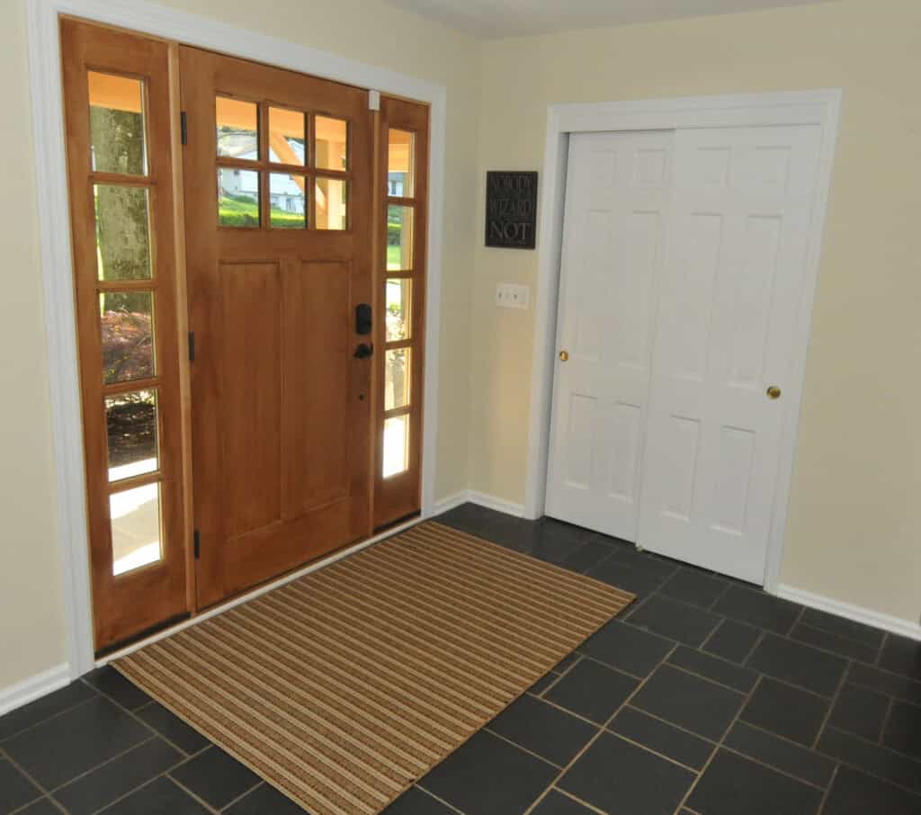 A bright entryway with a wooden front door featuring glass panels, white trim, a striped rug on dark tiled floor, and a set of white double closet doors.