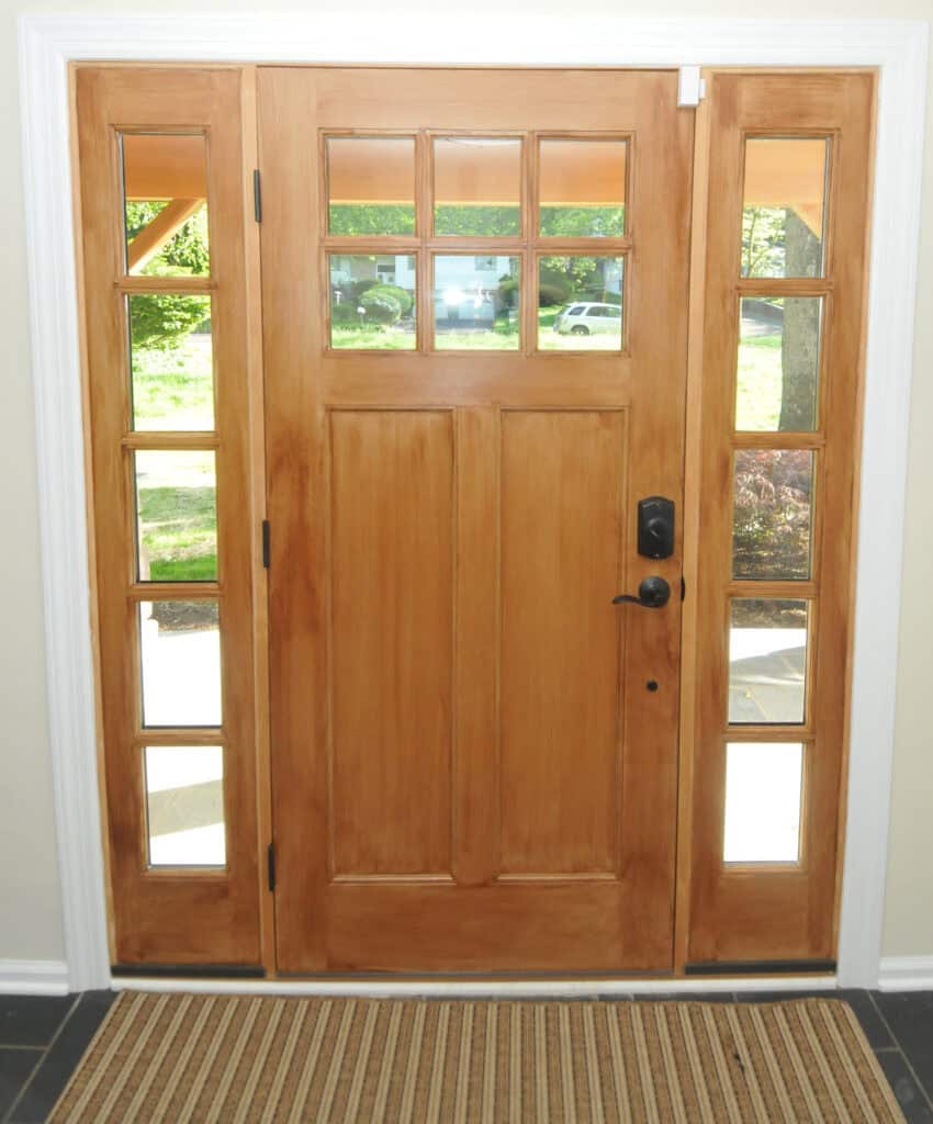 A wooden front door with rectangular panels, a black handle and lock, and decorative glass windows above and on both sides, allowing natural light to enter. A striped doormat lies in front of the door.
