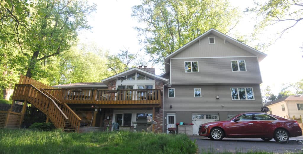A modern two-story house with beige siding, a wooden deck with stairs, and a red car parked in the driveway, surrounded by green trees and grass on a sunny day.