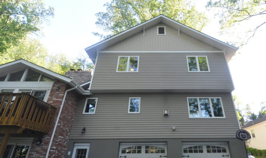 A three-story house with beige siding, white trim, and two garage doors. A wooden deck and brickwork are on the left side. Large windows reflect trees, and a basketball hoop is mounted above one garage door.