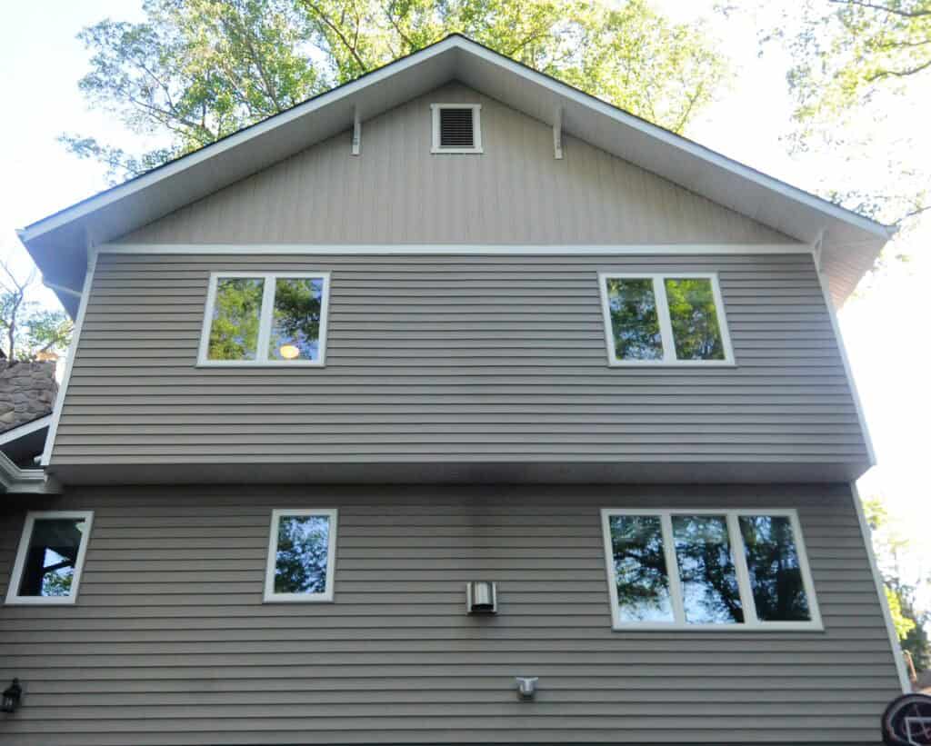 A two-story house with beige vinyl siding, several white-framed windows, a vent near the roof peak, and trees reflected in the glass and visible above the roofline.