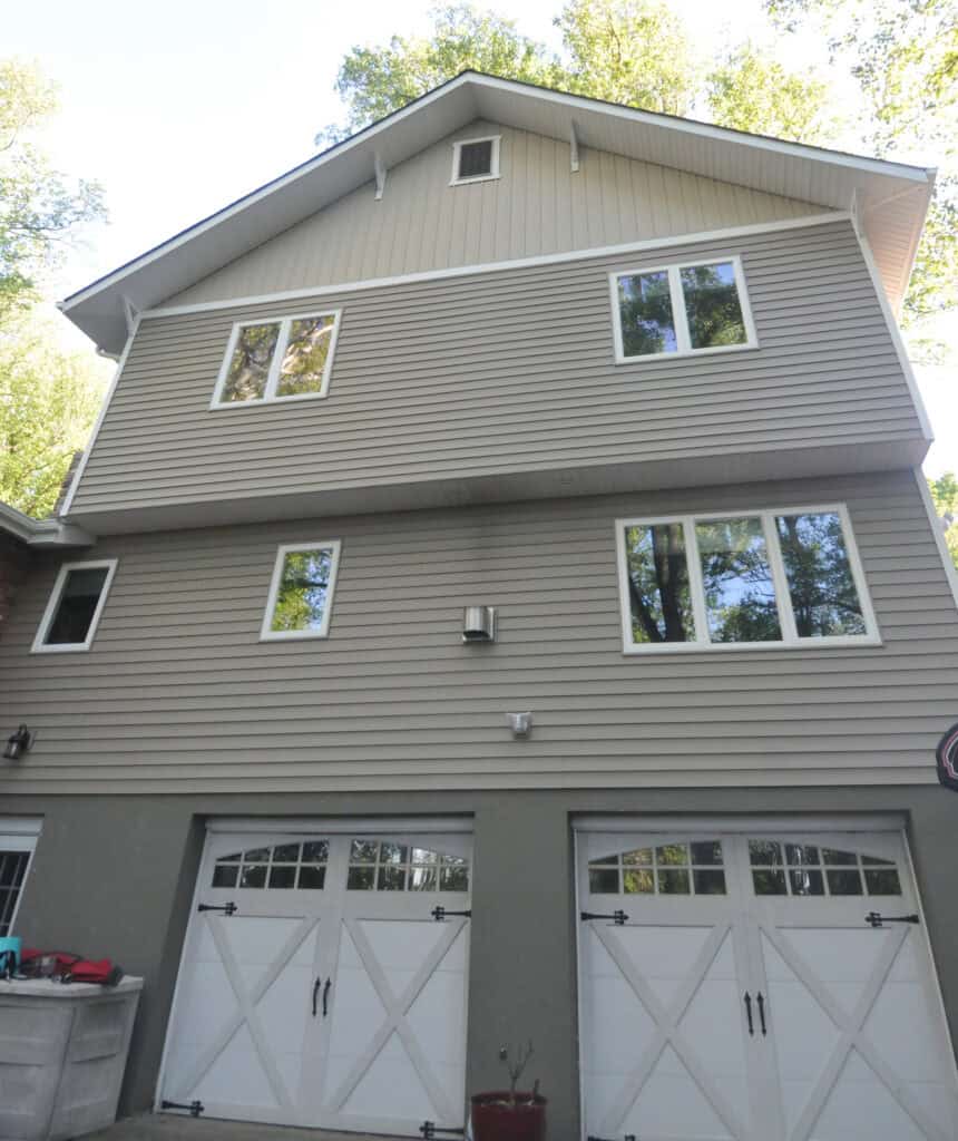 A two-story house with light brown siding, several white-framed windows, and two white garage doors with cross designs and windows at the top. Tall trees are visible in the background.