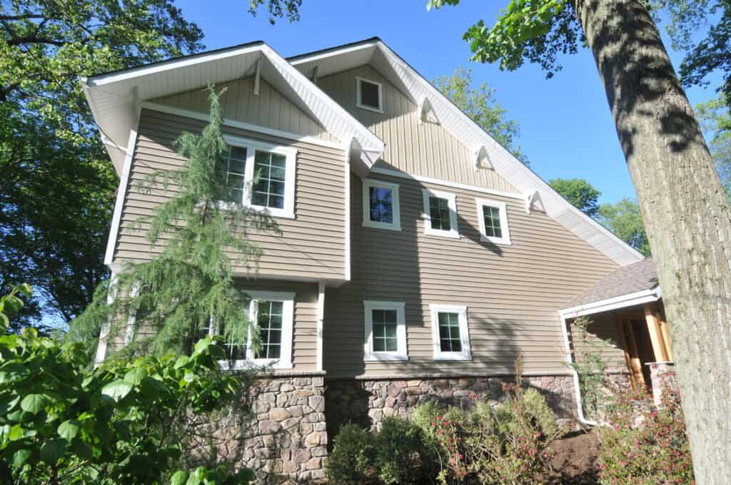 Tan house with stone foundation, white-trimmed windows, and gabled roof, surrounded by green trees and shrubs under a clear blue sky.