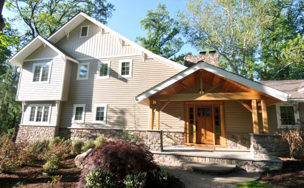 A two-story house with light tan siding, stone foundation, and a wooden covered front porch, surrounded by trees and landscaped shrubs on a sunny day.